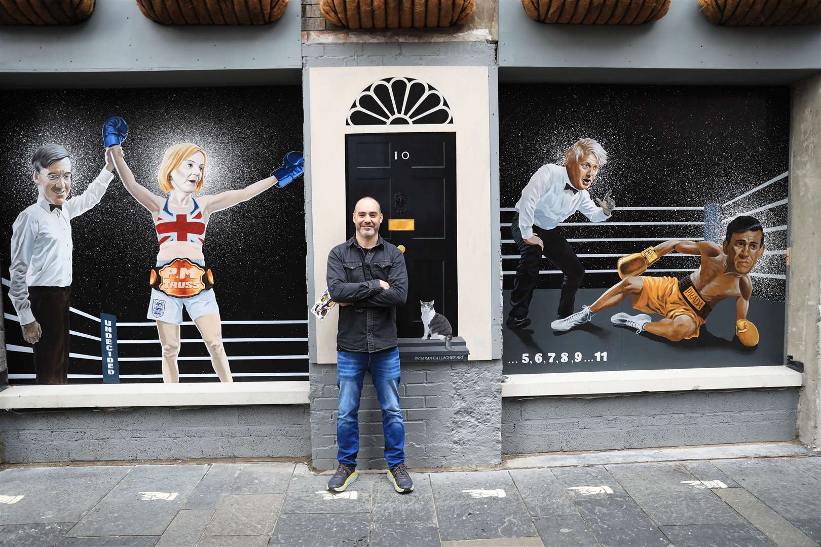 Artist Ciaran Gallagher beside his new mural depicting Liz Truss being declared a winner by Jacob Rees-Mogg after beating Rishi Sunak (Peter Morrison/PA)